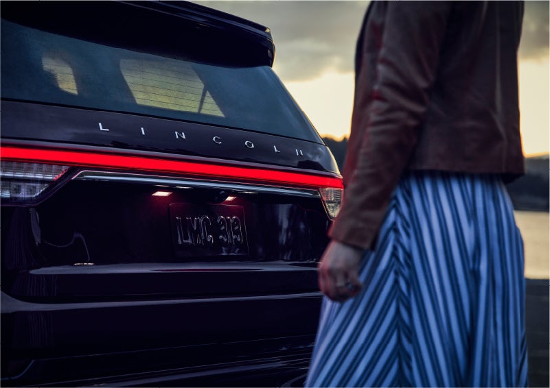 A person is shown near the rear of a 2023 Lincoln Aviator® SUV as the Lincoln Embrace illuminates the rear lights | Vision Lincoln in Wahpeton ND
