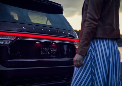 A person is shown near the rear of a 2024 Lincoln Aviator® SUV as the Lincoln Embrace illuminates the rear lights | Vision Lincoln in Wahpeton ND